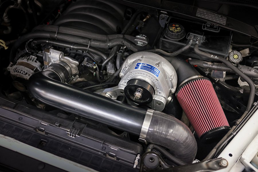 Redesigned ProCharger centrifugal supercharger system installed in a white GM 1500 pickup's engine bay, with a red cone air filter and black charge pipe